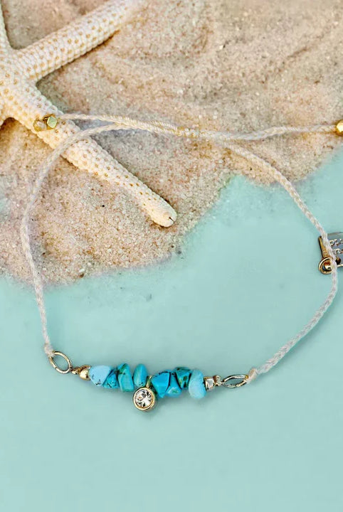 Pura Vida Teal Rock Bracelet with a Gold Charm Laying in Sand | Podos Boutique