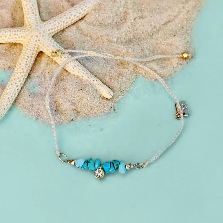 Pura Vida Teal Rock Bracelet with a Gold Charm Laying in Sand | Podos Boutique